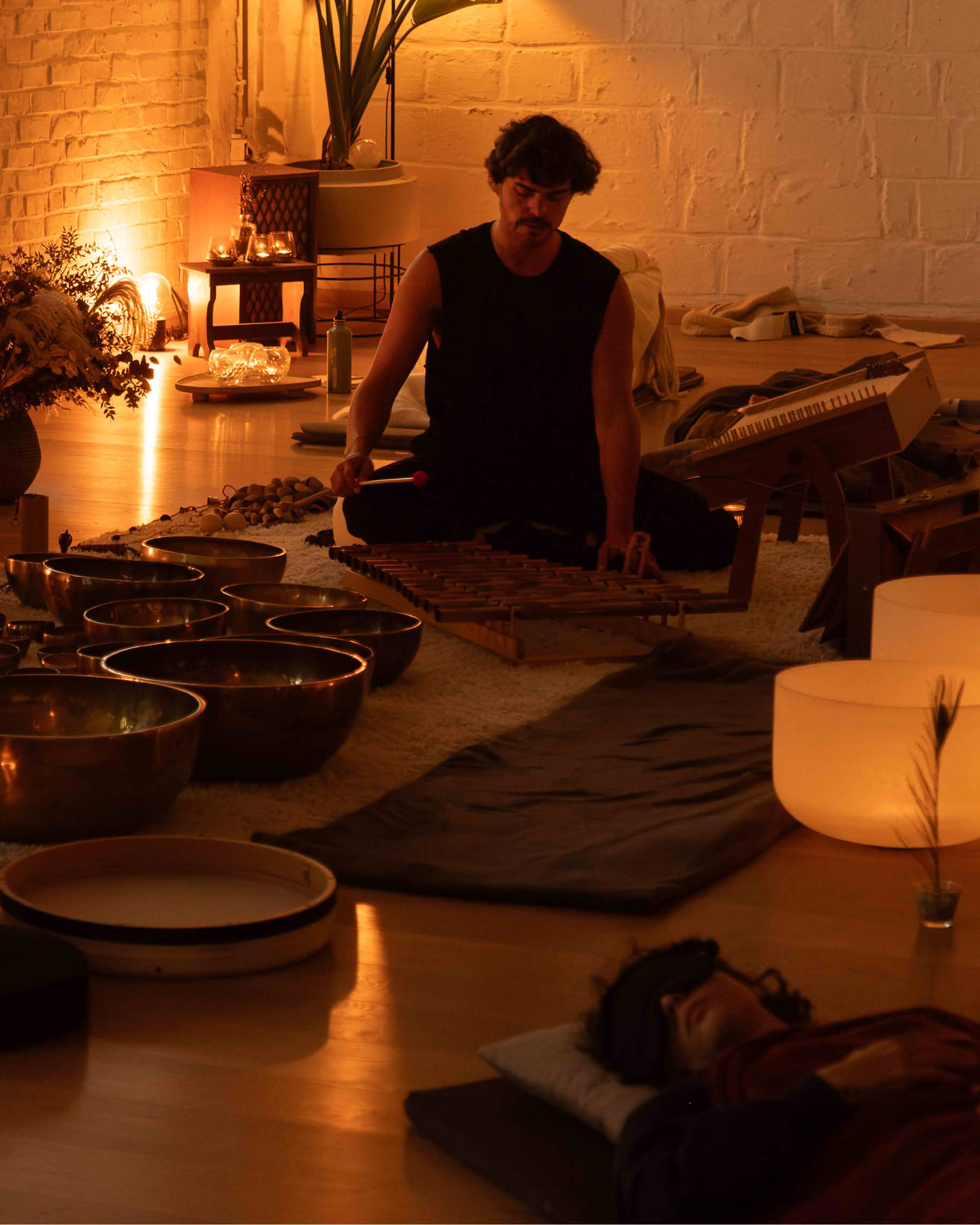 A sound healing practitioner sitting cross-legged amongst multiple Tibetan singing bowls, crystal bowls, and instruments in a dimly lit studio, leading a group meditative sound journey to help participants break through emotional blockages and release stress.