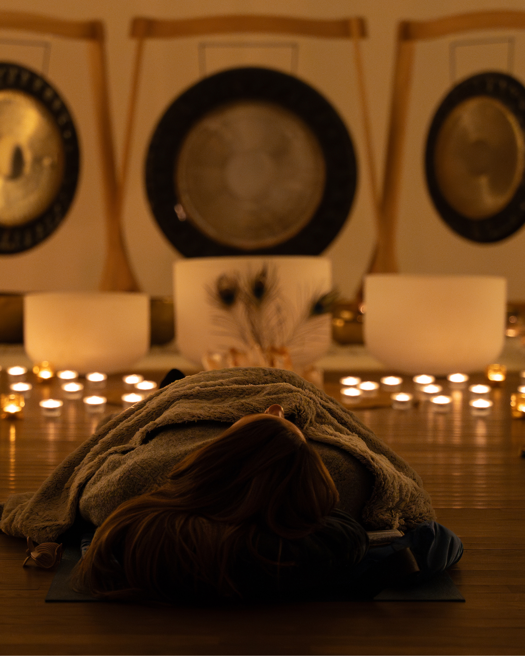 Relaxation and meditation: a woman relaxes on a mat in a sound therapy studio with lit candles, large gongs, and crystal singing bowls.